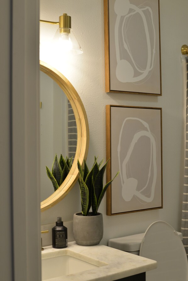 Un baño con un espejo redondo, una planta en maceta, una obra de arte abstracta enmarcada y un inodoro.