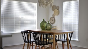 Un comedor con mesa de madera y sillas negras, persianas blancas y decoración en la pared.