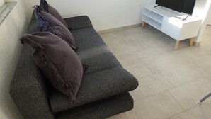 Ein graues Sofa mit zwei dunklen Kissen, eine weiße TV-Konsole mit einem Flachbildfernseher und ein gefliestes Parkett.