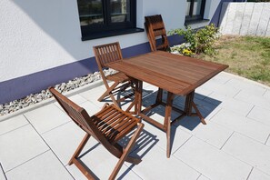 Una mesa de madera y cuatro sillas sobre un patio con baldosas, con una pared blanca y una ventana al fondo.