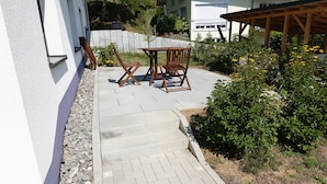 Eine Terrasse mit Holztisch und -stühlen, einem gepflasterten Weg und umgebender Bepflanzung.