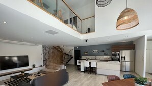 A modern living space with a large flat-screen TV, a sofa, a dining table with chairs, a kitchen area, and a staircase leading to an upper level.