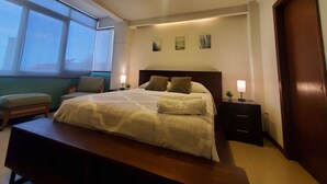 Un dormitorio moderno con una cama grande, mesitas de noche, un banco y vistas al cielo a través de una ventana.