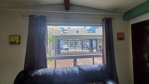 Una habitación con un sofá de cuero negro, una ventana con cortinas a rayas, vistas a una calle con coches y edificios, y una puerta cerrada.