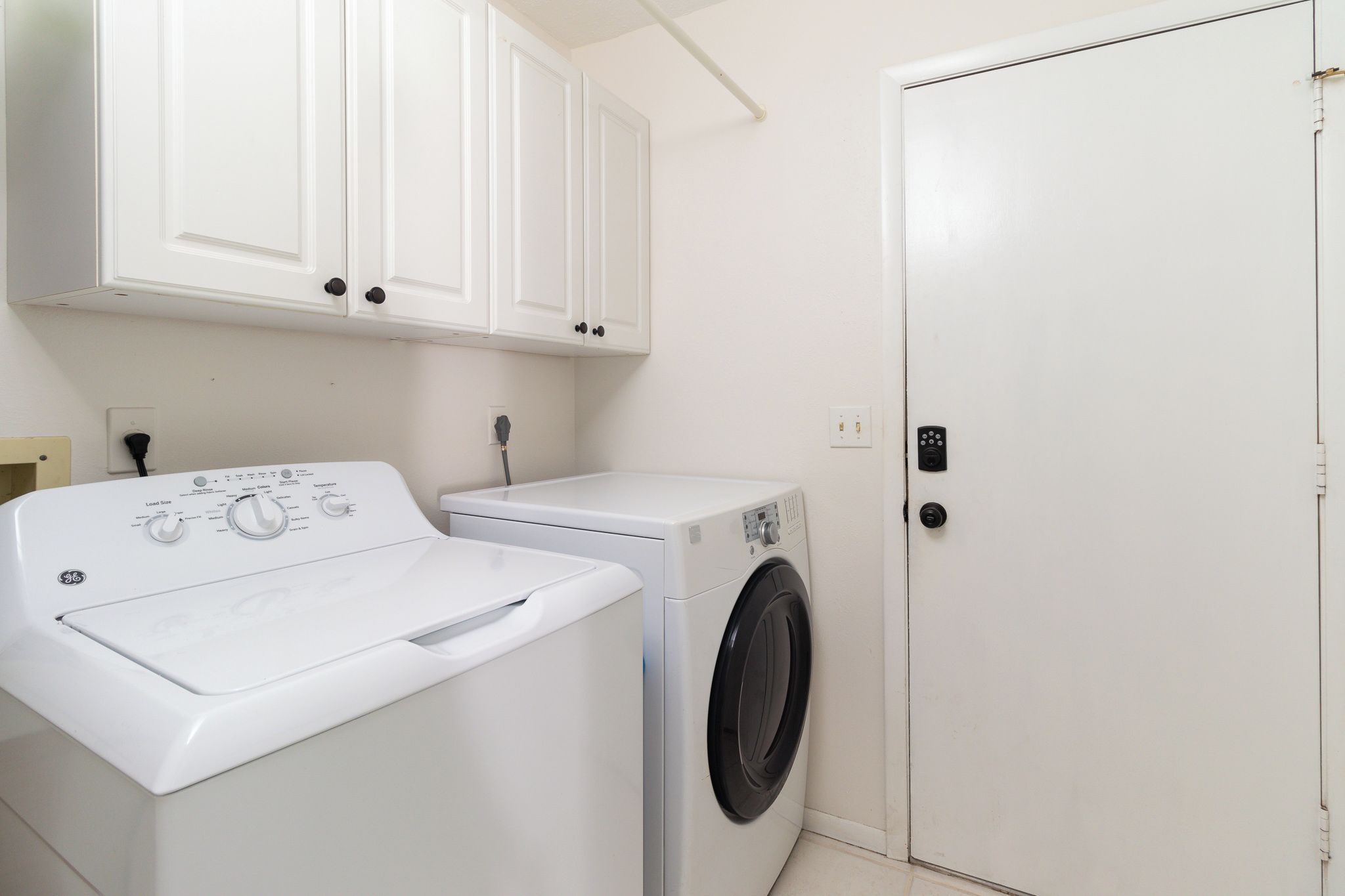 A laundry room with a front-loading washer and dryer, white cabinets above, and a white door with a keypad lock.