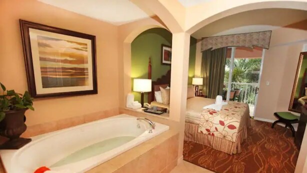 Habitación de hotel con una bañera grande, cama con ropa de cama floral, un escritorio pequeño y un balcón con vistas.