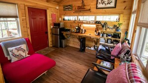 A cozy cabin interior with a red door, wooden walls, a kitchen area with a sink and stove, a red sofa, and a window with a view of trees. - Dreamy Treehouse Getaway ~Dog Friendly~Disc Golf (Copperhill)