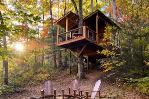 A treehouse with a wooden deck and a small porch, surrounded by a forest.