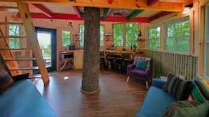 L’intérieur chaleureux d’un chalet avec un plancher en bois, un tronc d’arbre central, un coin repas et un espace salon comprenant un canapé et des fauteuils.