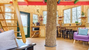 L’intérieur chaleureux d’un chalet avec un grand tronc d’arbre au centre, des murs en bois, un lit en mezzanine, un coin cuisine et un canapé violet.