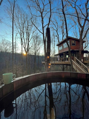 Una casa de madera con un puente de cristal sobre una superficie acuática reflectante, rodeada de árboles.