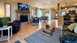 A spacious living room with a stone fireplace, leather sofas, a wooden coffee table, and a television mounted above the fireplace.