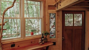 Interno di una baita rustica con soffitto in legno, una grande finestra con vista sugli alberi, una porta in legno e sgabelli rossi.