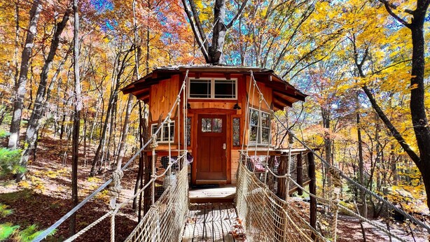 Una baita di legno con un ingresso su un ponte sospeso, circondata da alberi dai colori autunnali.