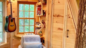 Un accogliente interno di una baita con soffitto in legno, una chitarra appesa al muro, una poltrona confortevole e un'ampia finestra.