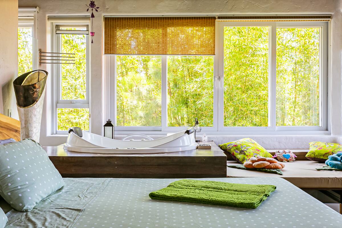 A room with a bed, a window with a view of greenery, a bathtub, and a window blind.