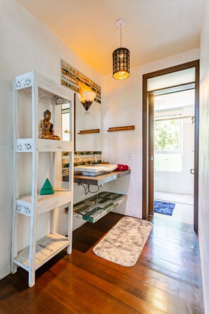 A bathroom with a white vanity, a sink, a mirror, a hanging light fixture, and a Buddha statue on a shelf. - Stupa EcoVila (Imbituba)