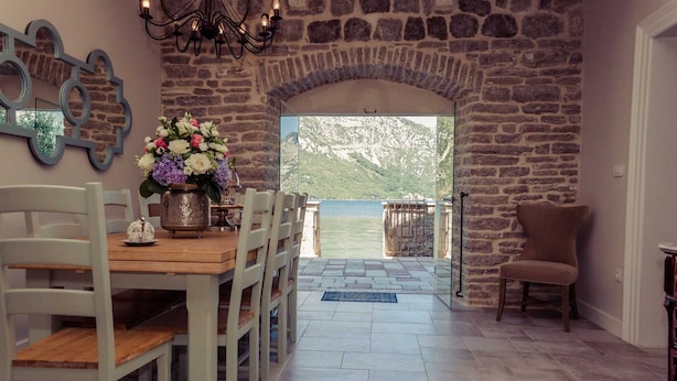 Zona pranzo con parete in pietra, tavolo in legno e sedie. Si vede un corpo d'acqua attraverso un arco aperto.