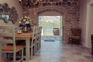 A dining area with a stone wall, a wooden table, and chairs. There is a view of a body of water through an open archway.