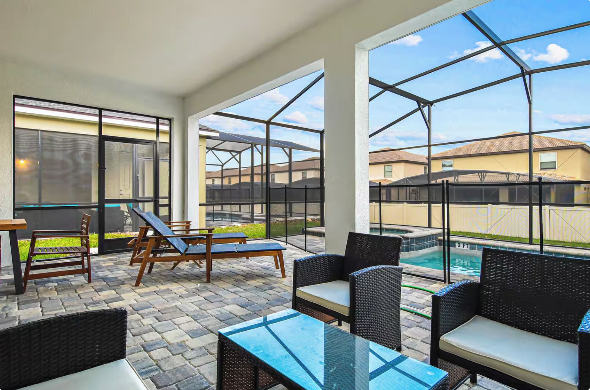 A covered patio with lounge chairs, a table, and a view of a swimming pool and houses.