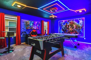 Una habitación con una mesa de fútbol de mesa, una televisión mostrando una película de superhéroes y un techo con luces de neón.