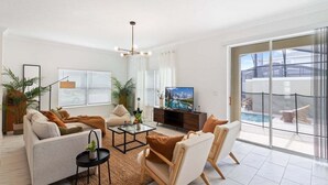 A modern living room with a sofa, armchairs, a coffee table, and a TV. There are potted plants and a view of a swimming pool through large windows. - 20% OFF Weekly Stay/ 5BR/Free Water Park (Kissimmee)