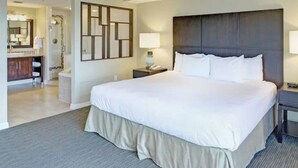 Habitación de hotel con cama grande, mesita de noche con lámpara, baño con espejo y lavabo, y zona de ducha.