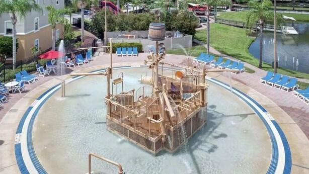 Parque acuático con una estructura central de juegos, zona de piscina circundante y sillas de descanso.
