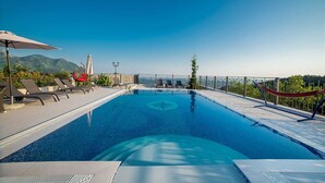 Una piscina a sfioro sul tetto con poltrone a sdraio e ombrelloni, circondata da una ringhiera metallica e vista sulle montagne.