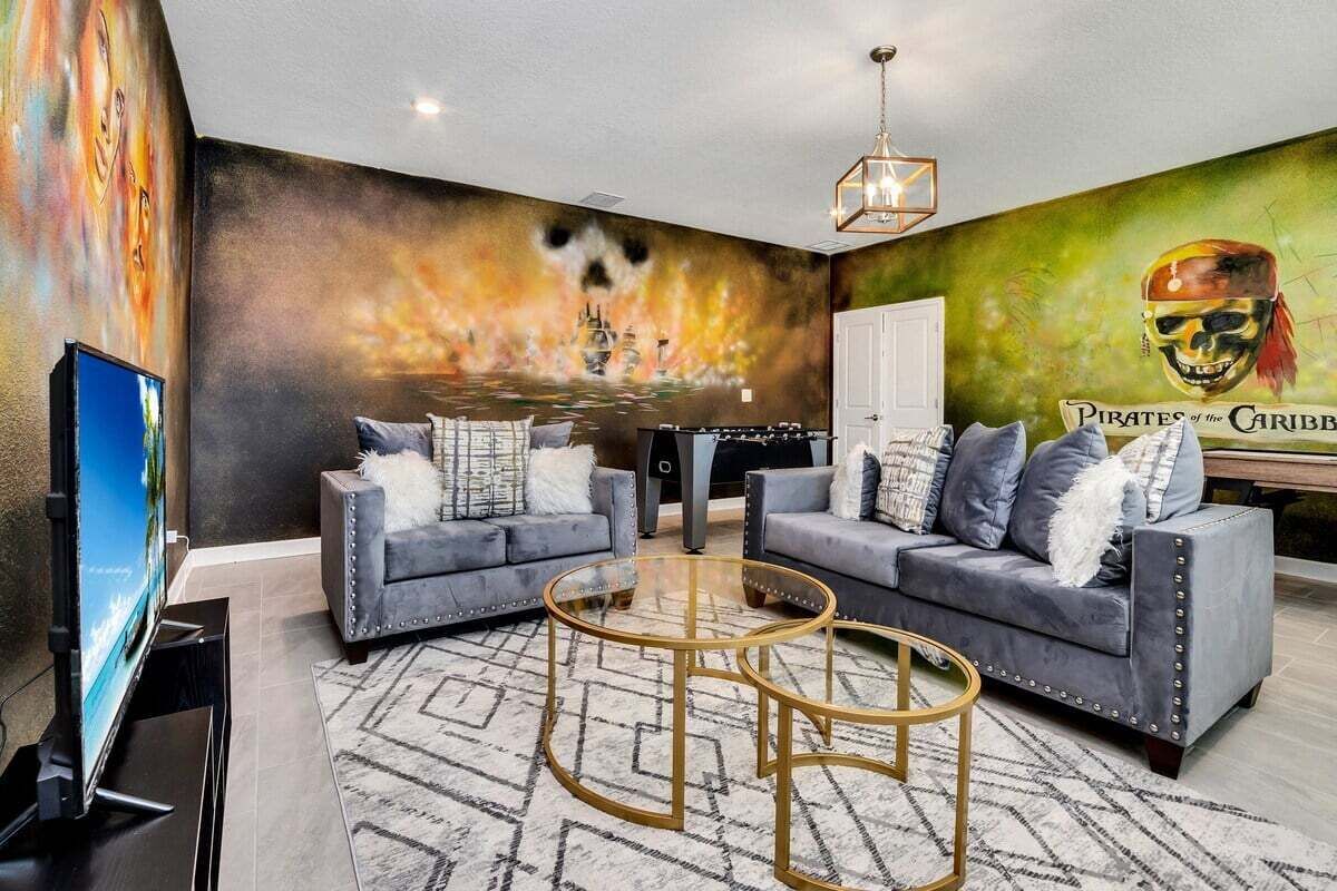 Een woonkamer met twee grijze banken, een glazen salontafel, een televisie en een muurschildering van een piratenschip.