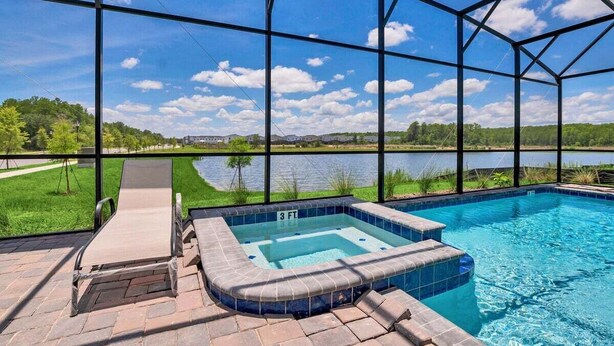 A swimming pool with a hot tub, surrounded by a glass enclosure, with a view of a lake and greenery. - Brand New Storey Lake 8BR Villa with Lake View (Kissimmee)