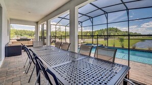 An outdoor patio with a metal table and chairs, a barbecue grill, and a view of a swimming pool and greenery. - Brand New Storey Lake 8BR Villa with Lake View (Kissimmee)