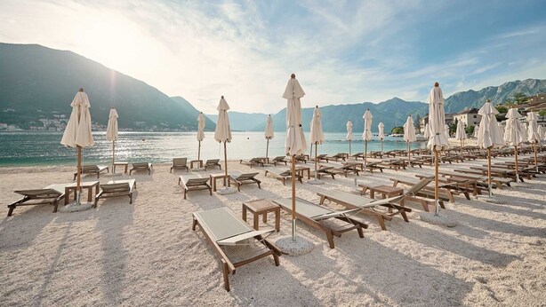 Une plage bordée de rangées de chaises longues et de parasols, une vue dégagée sur la mer et des montagnes en arrière-plan.