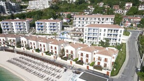 Un hotel frente al mar con varias edificaciones blancas, una playa con tumbonas y una carretera cercana.
