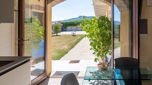 Una stanza con un tavolo in vetro, una sedia e vista su un giardino e montagne attraverso una porta aperta.