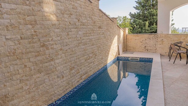 Une piscine dont le bord est revêtu de carreaux, entourée de murs en pierre et dotée d’un espace patio avec des chaises.