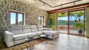 A modern living room with a stone wall, a grey sectional sofa, two black side tables, and a wooden floor. - Green Haven 2 Bedrooms (Budva)