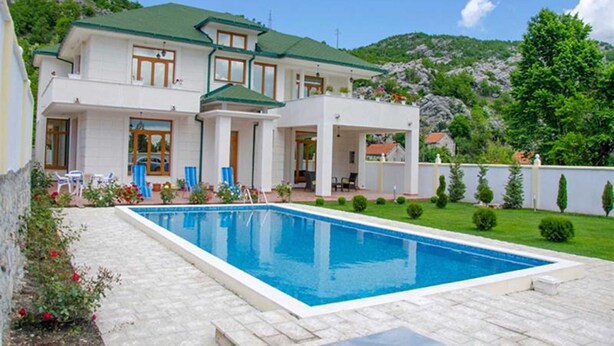 Uma casa de dois andares com piscina, cercada por um jardim bem cuidado e ao fundo, montanhas.