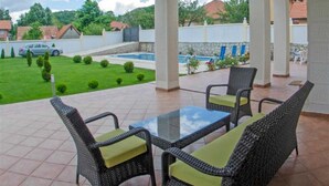 Um alpendre coberto com móveis de vime e uma mesa de vidro, com vista para uma piscina e um gramado verde.