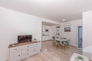 A modern living room with a TV, a dining area with a round table and chairs, and a kitchen with white cabinets.
