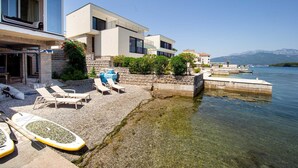 Un edificio moderno sulla costa, con vista diretta sul mare, una spiaggia di ciottoli con lettini e un pontile con barche.