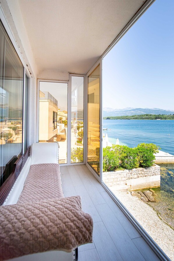 Ein Balkon mit Blick auf einen See und Berge, ein Bett mit rosa Decke und eine Glastür zu einem Außenbereich mit Tischen und Stühlen.