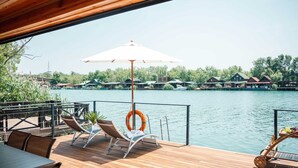 Una terraza de madera con sillones de descanso y una mesa, con vista a un lago tranquilo y casas al fondo.