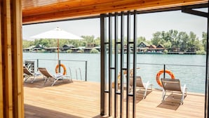Una terraza de madera con sillones de descanso y vistas a un lago con casas al fondo.