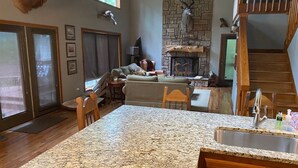 A spacious living room with a fireplace, a stone wall, and a kitchen area with granite countertops and wooden cabinets. - Beaver Lake Paradise with Bunkhouse (Springdale)