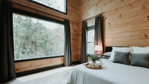 A wooden cabin bedroom with a large window, a bed with pillows, a bedside table with a lamp, and a small plant. -  Smoky Mnt Couples Cabin / Hot Tub / Secluded but close to local shops (Sevierville)