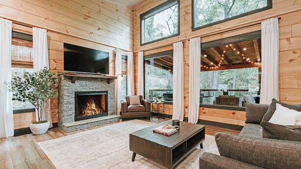 A cozy living room with a fireplace, comfortable seating, and large windows offering a view of the outdoors. -  Smoky Mnt Couples Cabin / Hot Tub / Secluded but close to local shops (Sevierville)