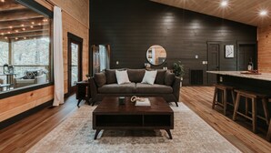 A modern living room with a dark wooden wall, a brown sofa with white cushions, a coffee table, and a large window with curtains. -  Smoky Mnt Couples Cabin / Hot Tub / Secluded but close to local shops (Sevierville)