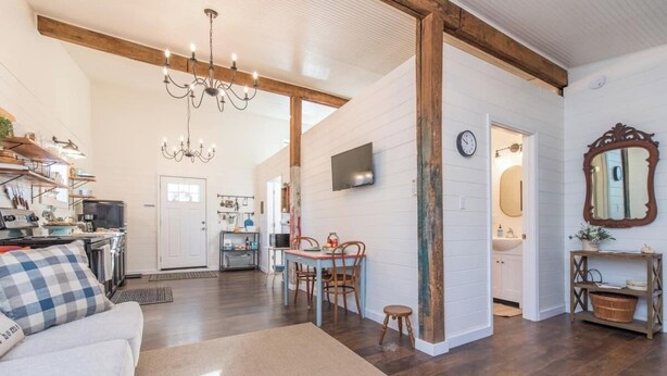 A spacious living room with a large chandelier, wooden beams, a dining area with a table and chairs, and a kitchen in the background.