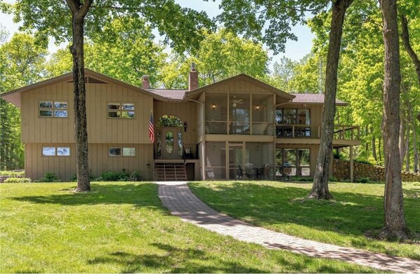 A two-story wooden house with a covered porch, a flag, and a paved driveway.